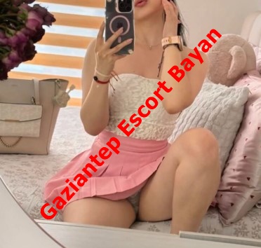Selfie of a woman in a white lace top and pink skirt posing on a bed, with a red 'Gaziantep Escort Bayan' overlay.